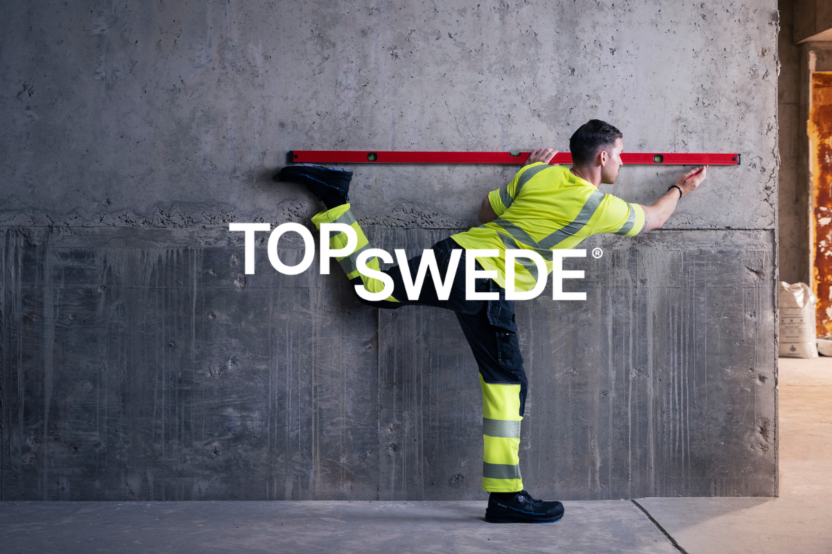 Byggarbetare i varselkläder och skyddsskor balanserar mot vägg med vattenpass. Klickbar länk till Top Swede-produkter